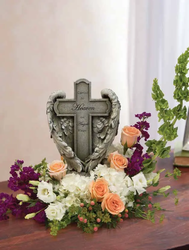 Angel wing cross statue surrounded by peach roses, white hydrangea and purple stock