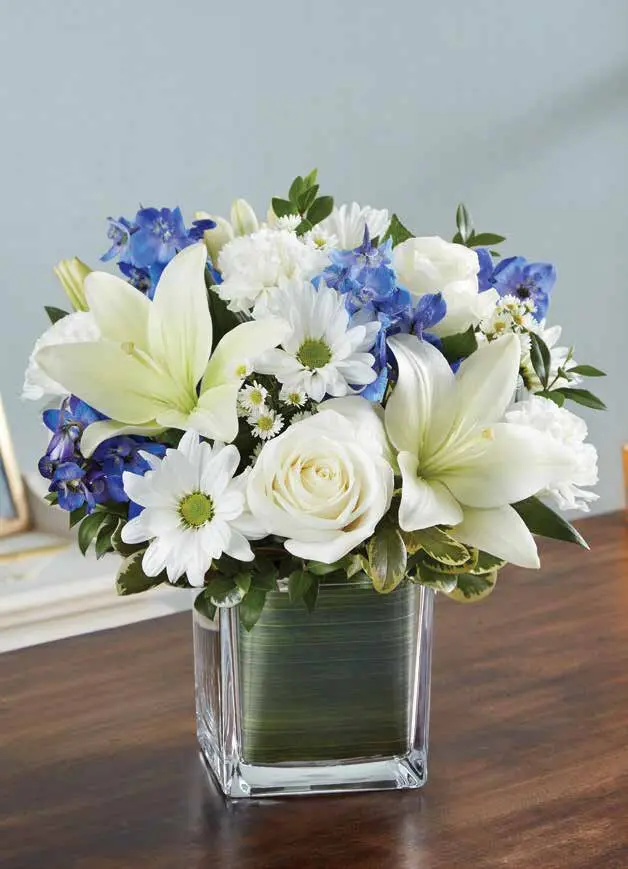 White lilies, roses and blue delphinium in a leaf-lined glass cube vase