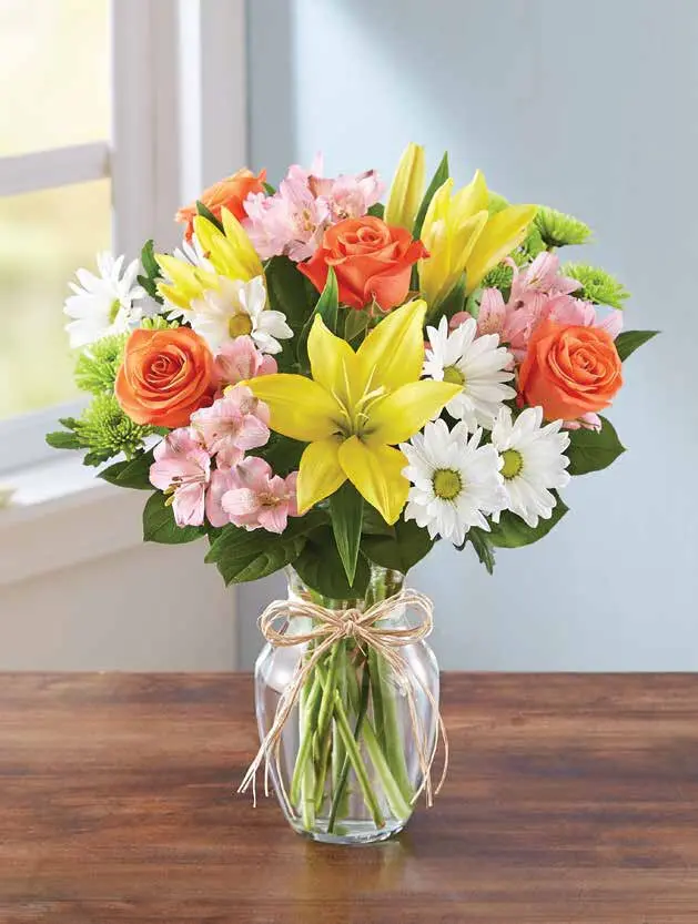 Yellow lilies, orange roses and pink alstroemeria in a clear vase with raffia bow