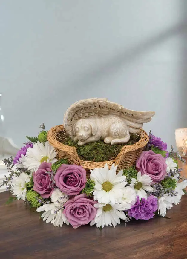 Sleeping puppy angel figurine in wicker bed ringed with mauve roses and daisies