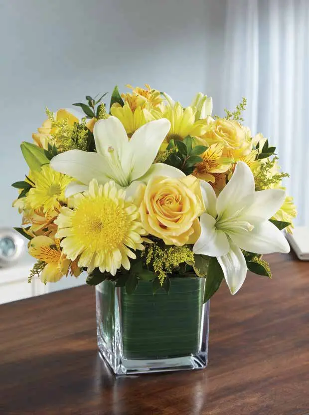White lilies, yellow roses and mums in a leaf-lined glass cube vase