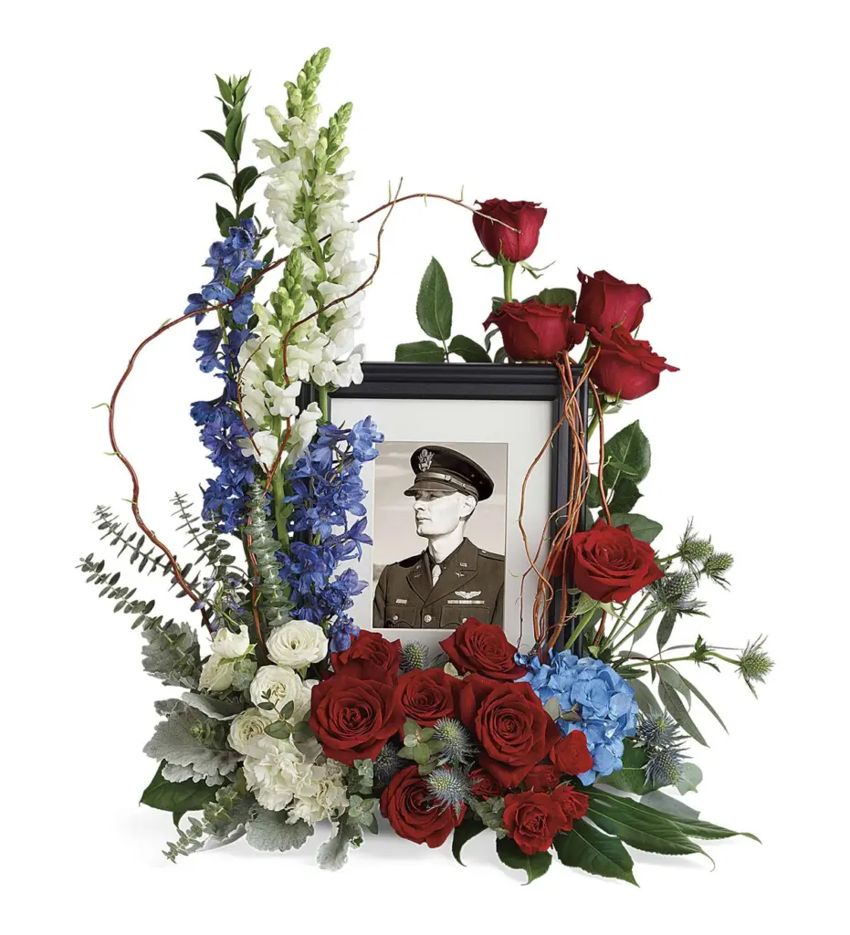 Red roses, blue delphinium and white snapdragons around a framed military portrait