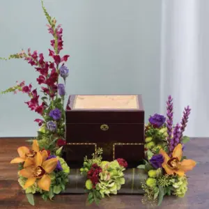 Urn surround of orange orchids, red snapdragons and purple lisianthus around a wooden urn