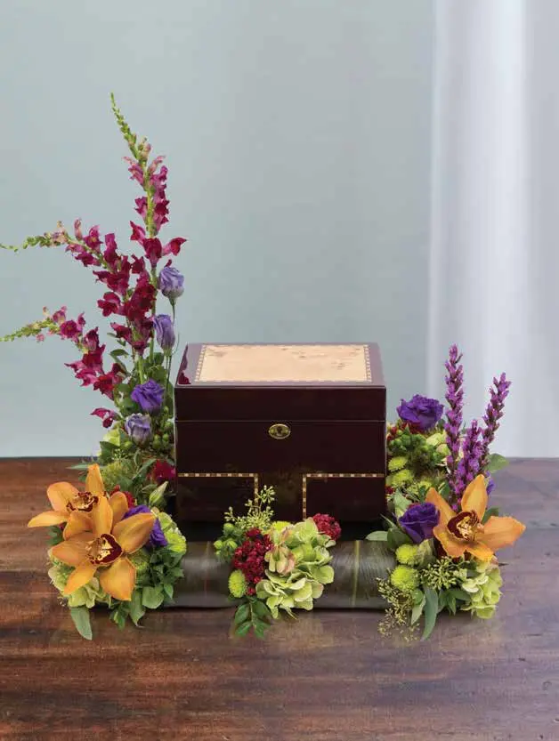 Urn surround of orange orchids, red snapdragons and purple lisianthus around a wooden urn